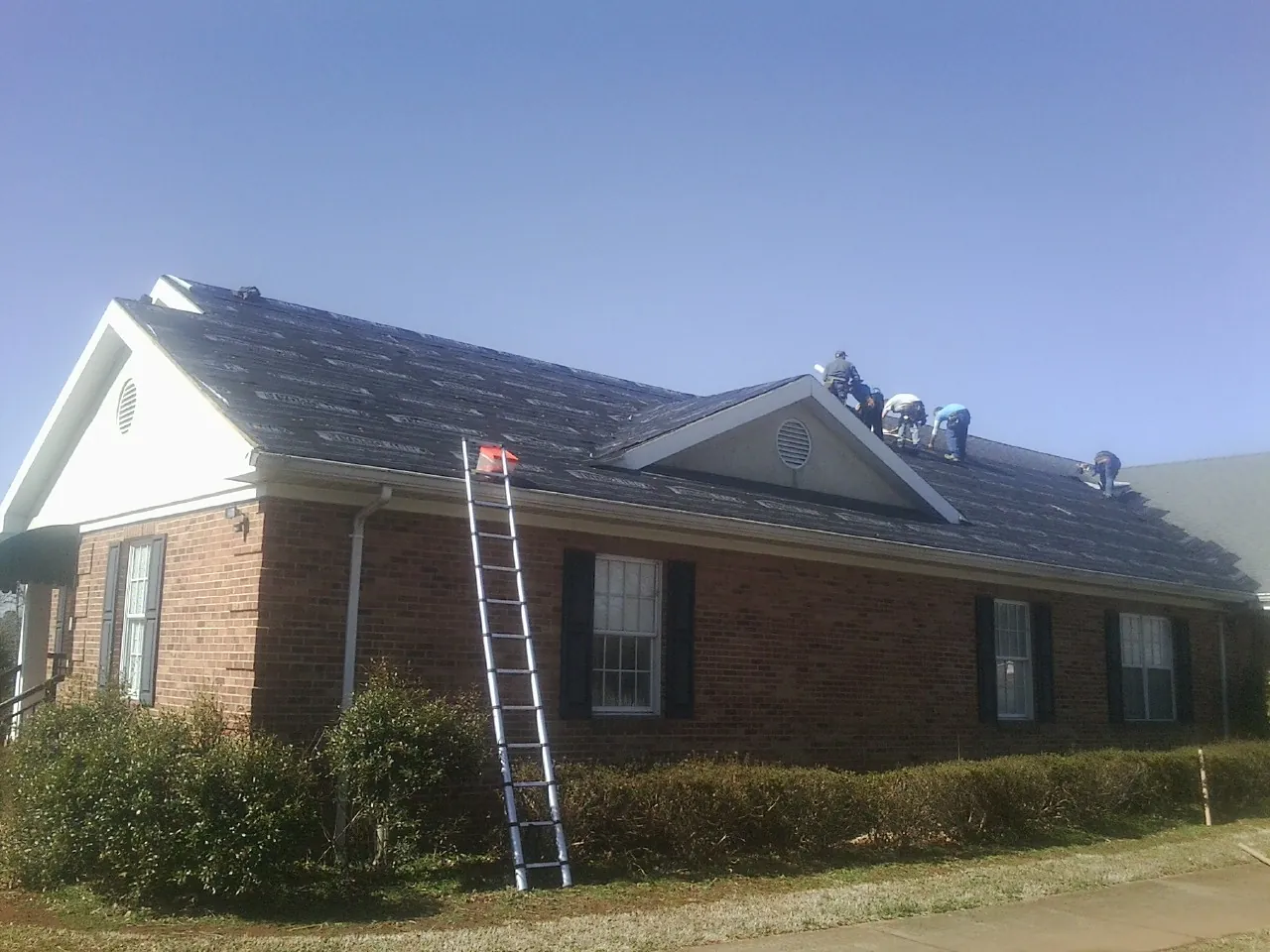 Crew tearing shingles off a brick ranch roof with a ladder set — Metal Roof Repair work by our crew in Bronson